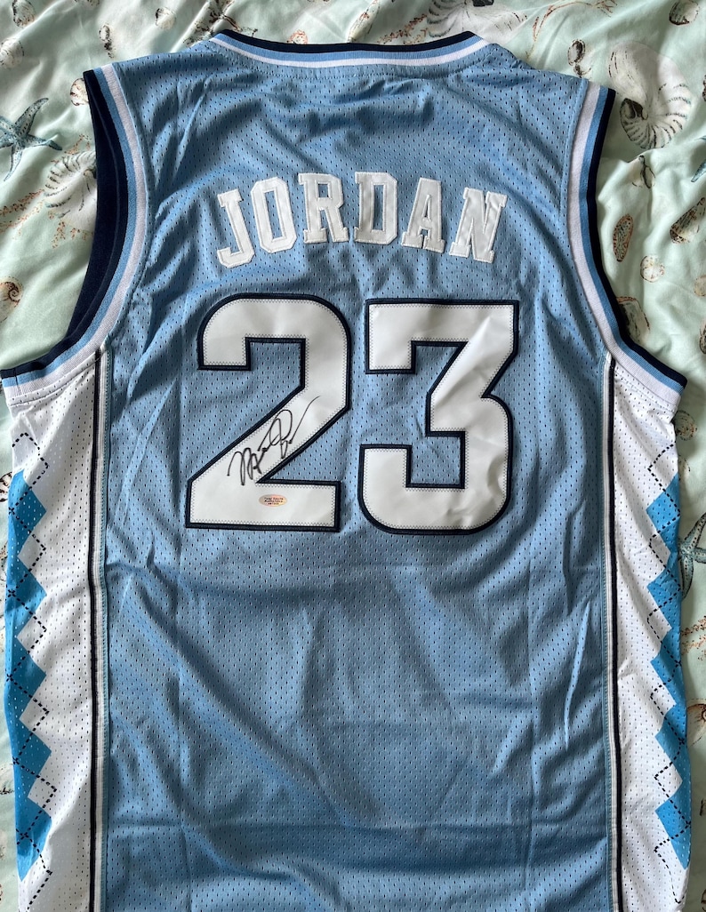 Może przedstawiać: Jasnoniebieski koszulka do koszyk&oacute;wki z napisem "JORDAN" i numerem "23" w kolorze białym. Koszulka ma podpis na numerze 23. Koszulka ma niebiesko-biały wz&oacute;r romb&oacute;w po bokach.