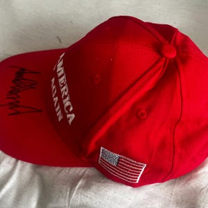 May include: A bright red baseball cap featuring the white embroidered text AMERICA AGAIN on the front panel. The curved brim displays a black signature, and a small rectangular patch of the United States flag is stitched onto the side panel.