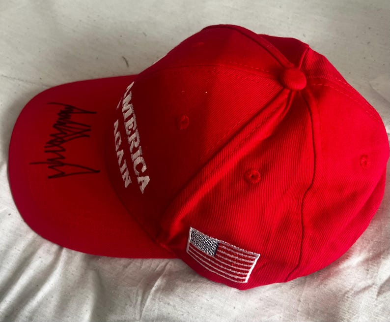 May include: A bright red baseball cap featuring the white embroidered text AMERICA AGAIN on the front panel. The curved brim displays a black signature, and a small rectangular patch of the United States flag is stitched onto the side panel.