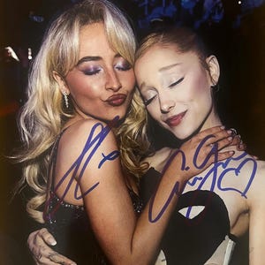 May include: A glossy photograph featuring two individuals with blonde and light brown hair embracing, adorned with blue ink signatures across the center. The bottom right corner displays a small white sticker with the text Total Sports Authentics 468578.