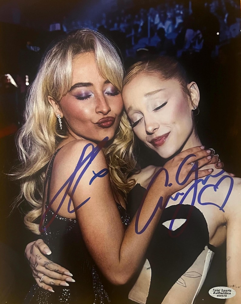 May include: A glossy photograph featuring two individuals with blonde and light brown hair embracing, adorned with blue ink signatures across the center. The bottom right corner displays a small white sticker with the text Total Sports Authentics 468578.