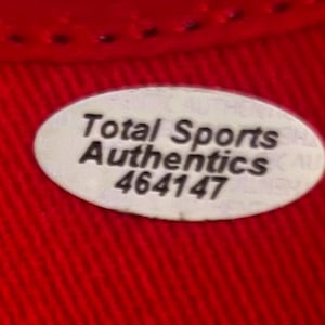 May include: An oval-shaped white sticker with a black border and black text that reads Total Sports Authentics 464147. The sticker is placed on a vibrant red fabric surface with a visible woven texture and a stitched seam along the top edge.