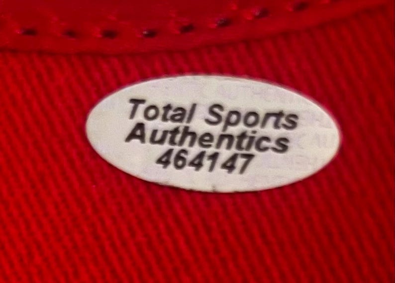 May include: An oval-shaped white sticker with a black border and black text that reads Total Sports Authentics 464147. The sticker is placed on a vibrant red fabric surface with a visible woven texture and a stitched seam along the top edge.
