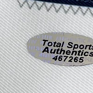 Może przedstawiać: Zbliżenie białej tkaniny z niebieską obw&oacute;dką i srebrną owalną naklejką. Na naklejce widnieje napis "Total Sports Authentics" i numer "467265".
