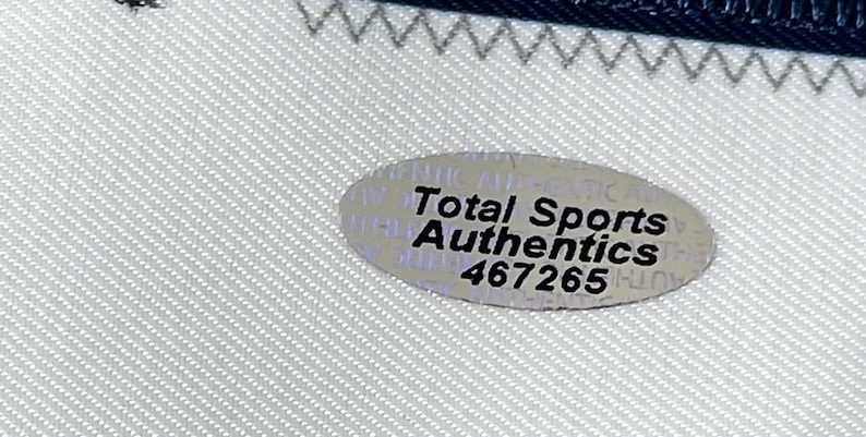 Może przedstawiać: Zbliżenie białej tkaniny z niebieską obw&oacute;dką i srebrną owalną naklejką. Na naklejce widnieje napis "Total Sports Authentics" i numer "467265".
