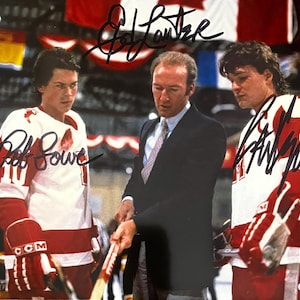 May include: A color photograph featuring three individuals on an ice rink, signed by Ed Lauter, Rob Lowe, and Patrick Swayze. Two individuals wear white and red hockey jerseys with gloves, while the central figure wears a dark suit and tie.