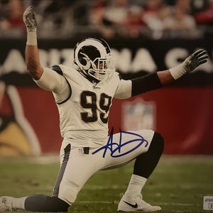 Puede incluir: Fotografía firmada de un jugador de fútbol americano con uniforme blanco y azul, número 99, arrodillado en un campo verde. El jugador lleva un casco con un diseño en blanco y negro. La firma es en tinta azul.