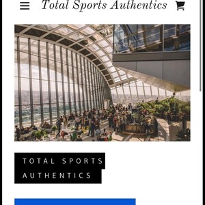 Może przedstawiać: Zrzut ekranu strony internetowej Total Sports Authentics. Obraz przedstawia duży, nowoczesny budynek ze szklanym dachem i wieloma ludźmi. Wyświetlany jest tekst "Total Sports Authentics" wraz z przyciskiem "Zweryfikuj autentyczność".