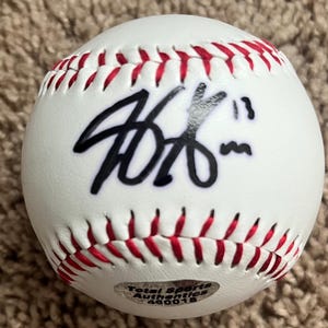 May include: A white baseball with red stitching. The ball features a black autograph and a Total Sports Authentics sticker. The baseball is a sports collectible.