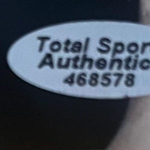 May include: An oval-shaped white sticker with a black border featuring the text Total Sports Authentics and the number 468578 printed in black. The sticker is positioned against a dark background, providing a clear view of the authentication label.
