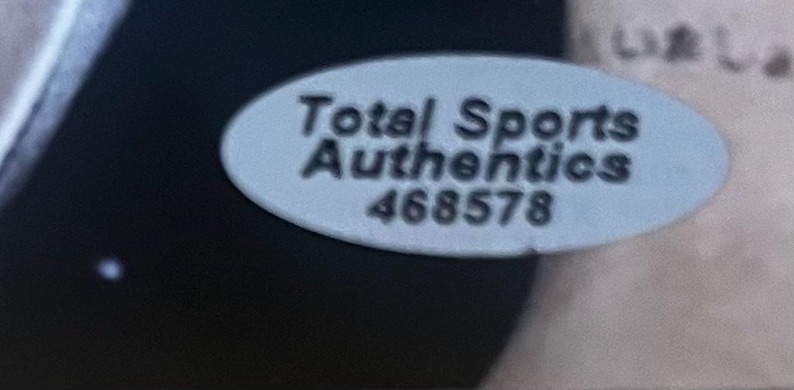 May include: An oval-shaped white sticker with a black border featuring the text Total Sports Authentics and the number 468578 printed in black. The sticker is positioned against a dark background, providing a clear view of the authentication label.