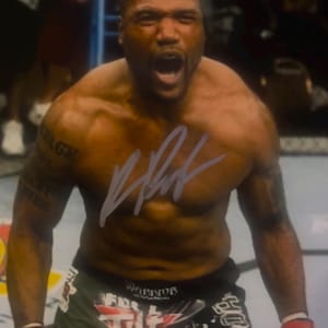 Puede incluir: Una fotografía de un luchador musculoso con la cabeza rapada, gritando con la boca abierta. El luchador viste pantalones cortos de combate negros y verdes con el texto Full Tilt Poker y Warrior, y guantes rojos y negros con una firma superpuesta.