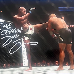 May include: Action shot of two athletes in a mixed martial arts match. One athlete is kicking the other. The image is signed "The Champ Jon Jones". The athletes are wearing athletic shorts and gloves. The background shows a crowd and the words "Tires" and "Premier Center".