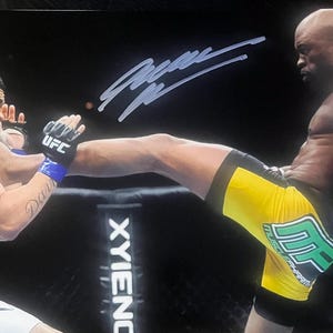 May include: A signed photograph of a mixed martial arts match featuring a fighter in yellow and green shorts delivering a front kick to the face of an opponent wearing black UFC gloves. The background shows a cage post with the text XYIENCE.