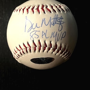 May include: A white baseball with red stitching featuring a blue ink autograph that reads Don Mattingly and 85 AL MVP. The ball includes a small, dark, oval-shaped authentication hologram sticker positioned near the bottom edge of the sphere.