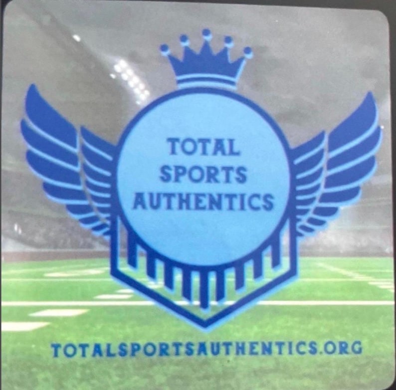 May include: A square sticker featuring a blue circular emblem with a crown on top and wings on the sides. The text TOTAL SPORTS AUTHENTICS is centered in the circle, and the website TOTALSPORTSAUTHENTICS.ORG is printed in blue at the bottom.