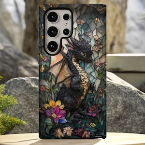 Op de afbeelding: Een telefoonhoesje met een glas-in-lood ontwerp met een zwarte draak, bloemen en vlinders. De draak heeft gedetailleerde vleugels en is omringd door kleurrijke bloemen. De hoes is geplaatst tegen een natuurlijke achtergrond met rotsen en groen.