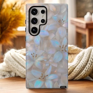 以下が含まれることがあります： 花柄のデザインのスマートフォンケース。ケースは、白と水色の花のパターンと虹色のアクセントが特徴です。携帯電話ケースは木製の表面にあります。