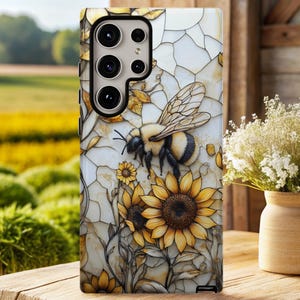 Op de afbeelding: Telefoonhoesje met een gedetailleerde illustratie van een hommel en zonnebloemen. Het ontwerp heeft een glas-in-lood effect met een witte en gouden achtergrond. De hoes wordt getoond op een houten oppervlak met een vage achtergrond van een veld en bloemen.