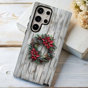 May include: A phone case featuring a Christmas wreath design. The case has a white wood grain background with a wreath of red poinsettias, green leaves, and berries. The phone case has a camera array.