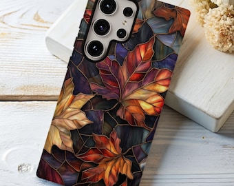 Puede incluir: Una funda para smartphone con un diseño de vidriera de hojas de otoño en tonos rojos, naranjas, amarillos y morados. La funda tiene un fondo oscuro y un acabado brillante, con un módulo de cámara en la parte superior.
