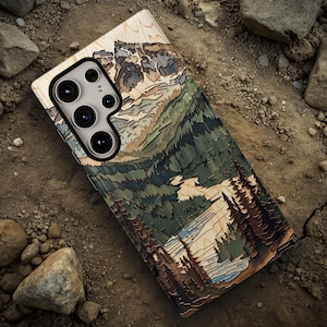 May include: A wooden phone case featuring a layered mountain and forest design in shades of brown, green, and beige. The case has a camera cutout and is lying on a dirt surface with rocks.