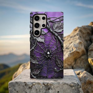 Puede incluir: Una funda de teléfono morada con un diseño de telaraña y una araña en el centro. La funda tiene un efecto texturizado en 3D con hojas y enredaderas plateadas. La funda del teléfono está sobre una superficie de piedra.
