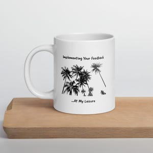 Puede incluir: Taza de cerámica blanca con un diseño de palmeras negras y el texto "Implementing Your Feedback ...At My Leisure". La taza está sobre una superficie de madera.