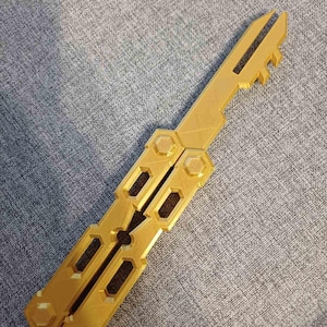 Può includere: Un coltello a farfalla da allenamento color oro caratterizzato da un design del manico scheletrato con ritagli esagonali e viti a perno. La lama è smussata e non affilata, con una distinta forma a chiave sulla punta, in materiale solido.