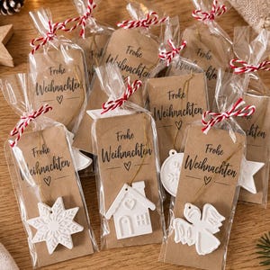 Könnte beinhalten: Eine Sammlung weißer Keramikanhänger in verschiedenen Formen wie Stern, Haus und Engel, einzeln verpackt in Klarsichtbeuteln mit einem braunen Papieranhänger, der mit dem Text Frohe Weihnachten und einem Herzsymbol bedruckt und mit rot-weißem Garn gebunden ist.