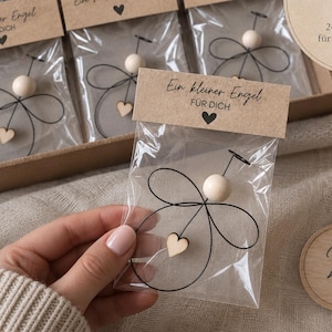 Könnte beinhalten: Ein Set aus kleinen Engel-Anhängern, verpackt in transparenten Kunststoffbeuteln mit braunen Papier-Etiketten. Jeder Engel besteht aus schwarzem Draht, einer hellen Holzperle als Kopf und einem kleinen Holzherz. Die Etiketten tragen deutschen Text.