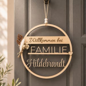 Könnte beinhalten: Ein rundes Holzschild mit der Aufschrift Willkommen bei FAMILIE Hildebrandt. Das Schild ist an einer Jutekordel mit zwei Holzperlen aufgehängt und auf der linken Seite mit einem kleinen Arrangement aus getrockneten weißen und braunen Blumen verziert.