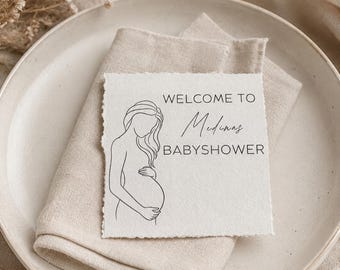 Könnte beinhalten: Eine quadratische, cremeweiße Karte mit Büttenrand, die eine schwarze Strichzeichnung einer schwangeren Figur und den gedruckten Text WELCOME TO Medina's BABYSHOWER zeigt. Die Karte liegt auf einer gefalteten beigen Leinen-Serviette auf einem Keramikteller.
