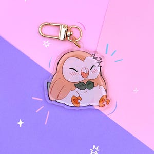 May include: A clear acrylic keychain featuring a cartoon owl with light brown feathers, a white belly, and a dark green bowtie. The owl has closed eyes, a smiling beak, and orange feet. A gold-toned metal lobster clasp is attached to the top.