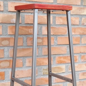 Op de afbeelding: Een metalen barstoel met een rode houten zitting. De stoel heeft een eenvoudig, industrieel ontwerp met een rechthoekige zitting en vier poten.