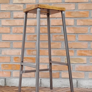 Op de afbeelding: Een metalen barstoel met een houten zitting. De stoel heeft een eenvoudig ontwerp met vier poten en een dwarsbalk voor ondersteuning. De stoel staat op een betegelde vloer.