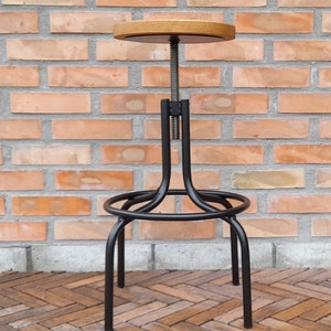 Puede incluir: Un taburete de bar de metal negro con un asiento de madera. El taburete tiene un diseño sencillo con un asiento redondo y tres patas. El taburete es ajustable en altura.