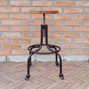 Op de afbeelding: Zwarte metalen barstoel met een houten zitting en wielen. De stoel heeft een eenvoudig ontwerp met een ronde zitting en een metalen frame.