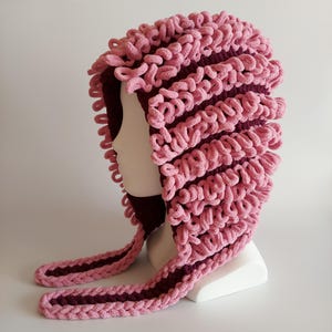 May include: A crochet hood featuring alternating stripes of soft pink looped yarn and solid dark maroon yarn. The design includes long, dangling side panels with a braided edge, creating a textured, layered appearance that covers the head and neck.