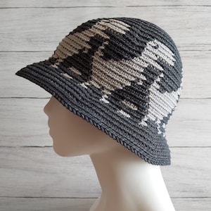 Puede incluir: Un sombrero de pescador de ganchillo gris oscuro y gris claro con un ala pequeña. El sombrero presenta un patrón geométrico en tonos grises alternos. El sombrero se muestra en una cabeza de maniquí sobre un fondo de madera blanca.