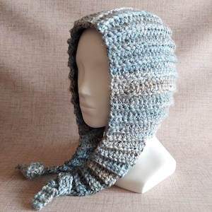 May include: A handmade crochet hood in shades of blue, gray, and white, displayed on a mannequin head. The hood features a ribbed design and long ties with decorative knots at the ends. The yarn has a textured appearance.