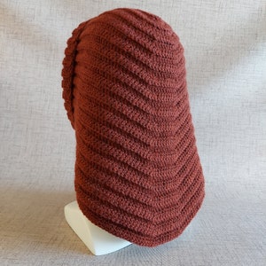 May include: A rust-coloured crocheted hat with a unique, textured design. The hat is made with a thick yarn and has a slouchy fit.