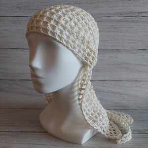 May include: Off-white crochet head covering with a net-like pattern. The head covering is displayed on a white mannequin head. The head covering has a long, flowing tail that hangs down the back. The crochet work appears to be made of a soft, lightweight yarn.