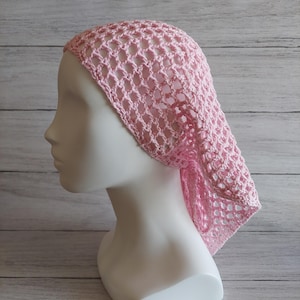 May include: A pale pink crochet head covering with an open, net-like weave. The head covering is displayed on a white mannequin head, highlighting the delicate design. The crochet pattern creates a light and airy appearance.