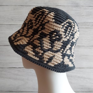 May include: A black and beige crocheted bucket hat with a geometric pattern. The hat is on a white mannequin head.