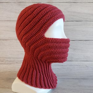Puede incluir: Una balaclava de punto de ganchillo roja con un largo cuello que la cubre. La balaclava tiene un agujero para los ojos y la nariz.