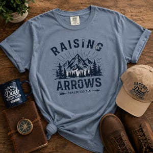 Puede incluir: Una camiseta azul de manga corta con un gráfico azul oscuro de montañas, pinos y el texto "RAISING ARROWS" con "PSALM 127:3-5" debajo. La camiseta se muestra junto a una taza azul oscuro, una gorra de béisbol beige, un diario y botas.