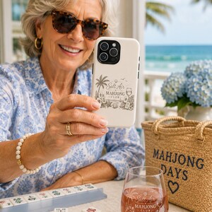 May include: A cream-colored smartphone case and a clear stemless wine glass, both featuring a black graphic print that reads Salt Air Mahjong Club, accompanied by illustrations of palm trees, seashells, and mahjong tiles.