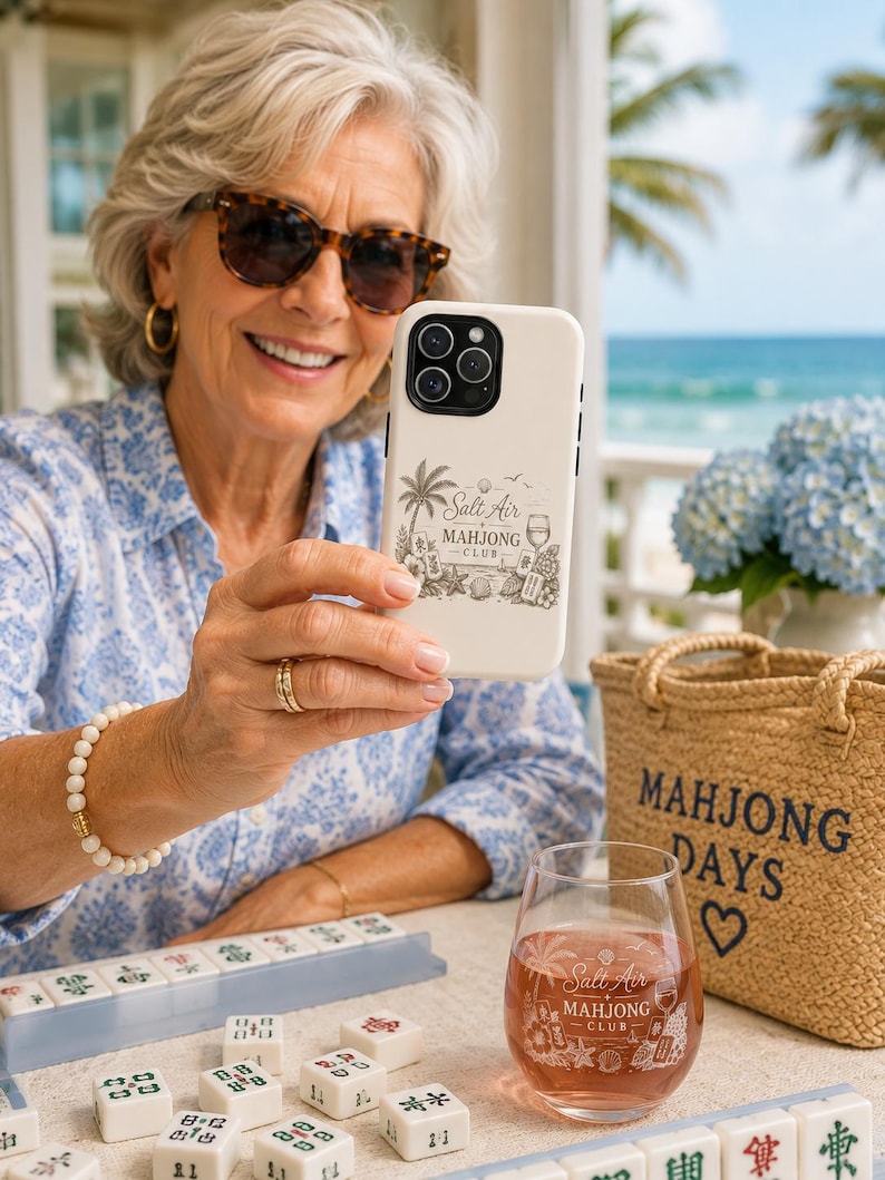 May include: A cream-colored smartphone case and a clear stemless wine glass, both featuring a black graphic print that reads Salt Air Mahjong Club, accompanied by illustrations of palm trees, seashells, and mahjong tiles.