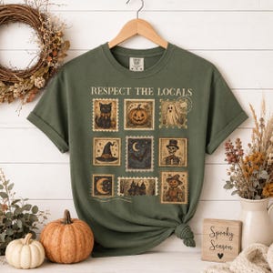 May include: An olive green short-sleeve t-shirt featuring the text RESPECT THE LOCALS above a grid of nine vintage-style postage stamp illustrations depicting a black cat, pumpkin, ghost, witch hat, bat, skeleton, moon, house, and scarecrow.